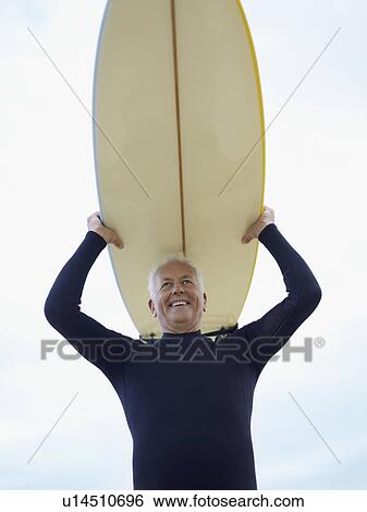 Stock Photograph - Senior man carrying surfboard on head, (low angle view). Fotosearch