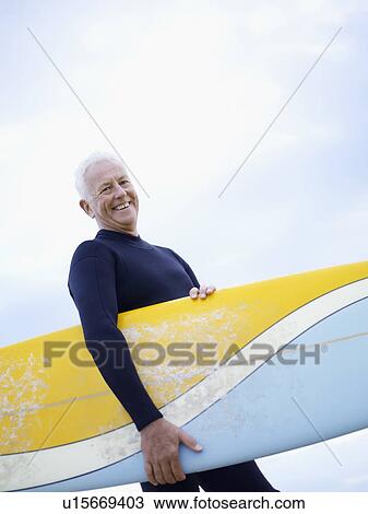 Stock Image - Senior man carrying surfboard, smiling. Fotosearch