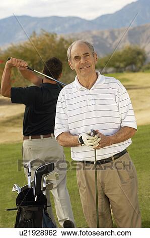 Stock Image - Senior man holding golf club, standing on golf course. Fotosearch