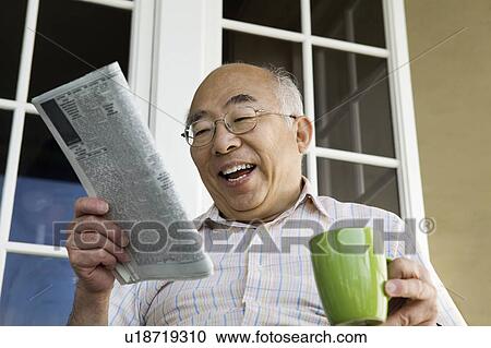 Stock Image - Senior man reading newspaper on porch, laughing, (low angle view). Fotosearch