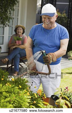 Stock Image - Senior man watering plants in garden, wife in background. Fotosearch
