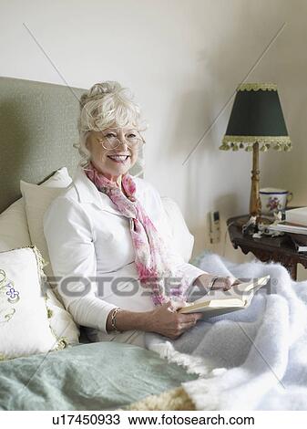 Stock Image - Senior woman sitting on bed, reading book. Fotosearch