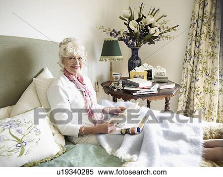 Stock Photography - Senior woman sitting on bed, reading book. Fotosearch