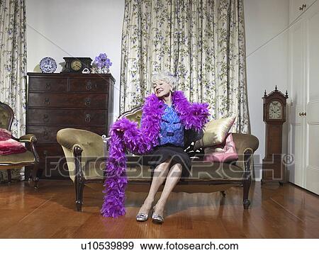 Stock Photo - Senior woman wearing feather boa sitting on love seat in living room, (portrait). Fotosearch