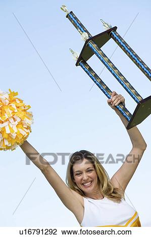 Stock Image - Smiling Cheerleader holding trophy, (portrait), (low angle view). Fotosearch