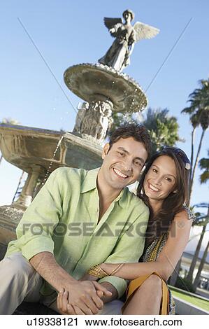 Stock Image - Smiling Couple, arms around, sitting in Front of Fountain. Fotosearch