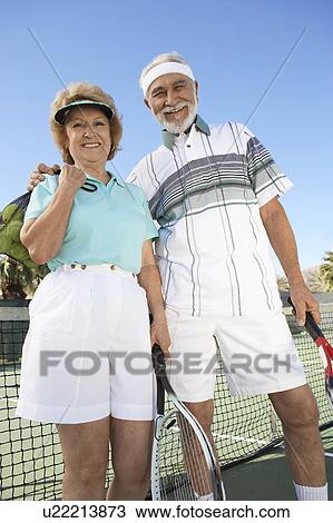 Stock Image - Smiling Couple standing side by side on Tennis Court, holding rackets. Fotosearch