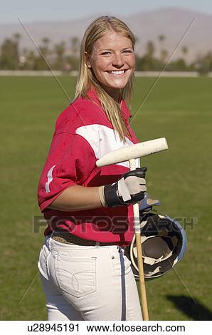 Stock Image - Smiling Female Polo Player standing on polo field holding polo stick and helmet, side view. Fotosearch