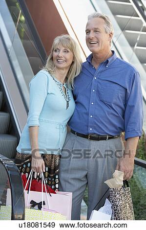 Smiling middle-aged Couple standing, arms around each other near escalator on Shopping Trip View Large Photo Image Stock Photo - Smiling middle-aged Couple standing, arms around each other near escalator on Shopping Trip. Fotosearch