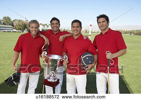 Stock Image - Smiling Polo Players standing on polo field, holding equipment and trophy. Fotosearch