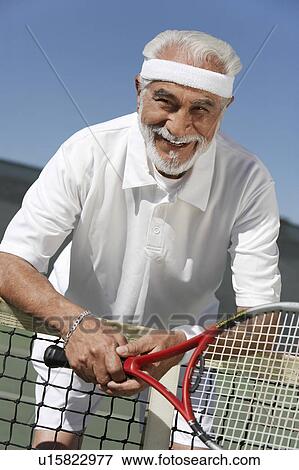 Stock Photo - Smiling senior Man leaning on tennis net, holding tennis racket. Fotosearch
