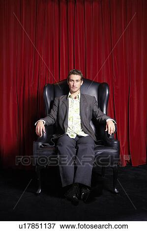 Stock Photo - Stylishly dressed Man sitting in Wingback Chair in front of curtain. Fotosearch