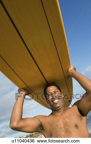 Stock Photograph - Surfer carrying surfboard on head at beach, (portrait). Fotosearch