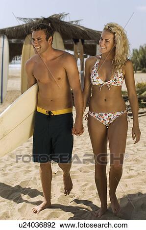 Stock Image - Surfer couple holding hands, carrying surfboard, walking on beach. Fotosearch