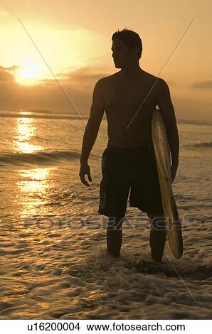 Picture - Surfer standing in surf, holding surfboard, at sunset. Fotosearch