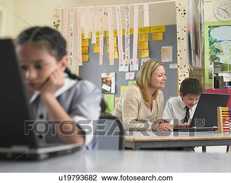 Stock Image - Teacher sitting with student using laptop in classroom. Fotosearch