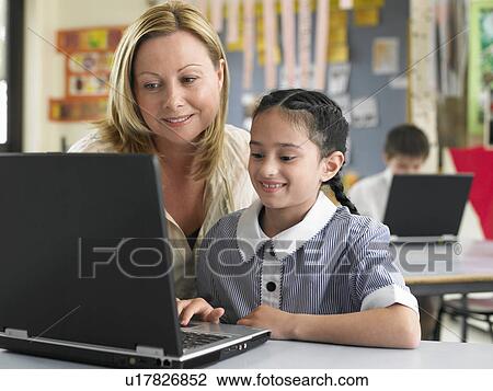 Stock Image - Teaching sitting with elementary schoolgirl using laptop in classroom. Fotosearch