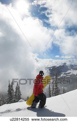 Stock Photography - Teenage boy (16-19) holding snowboard, hiking up snow covered slope, full length. Fotosearch