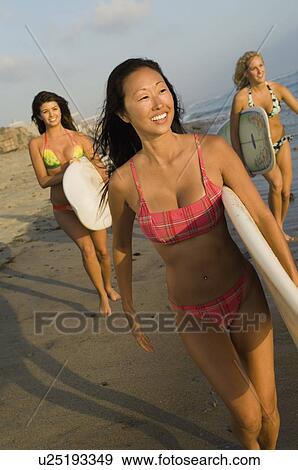 Stock Photo - Three female surfers carrying surfboards, walking on beach. Fotosearch