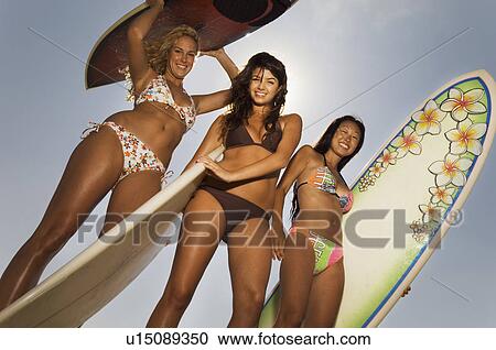 Stock Image - Three female surfers holding surfboards outdoors, low angle view. Fotosearch