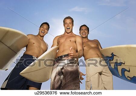 Stock Photograph - Three surfers standing holding surfboards outdoors, laughing, low angle view. Fotosearch