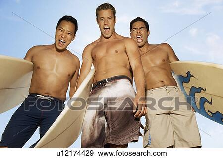 Stock Photo - Three surfers standing with surfboards, making faces, outdoors, low angle view. Fotosearch