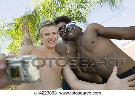 Stock Image - Three young men taking photo of themselves, low angle view. Fotosearch