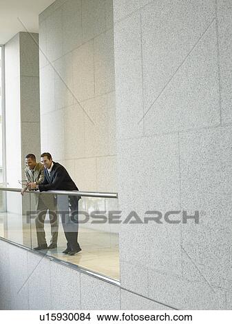 Picture - Two businessmen leaning on railing in office building. Fotosearch