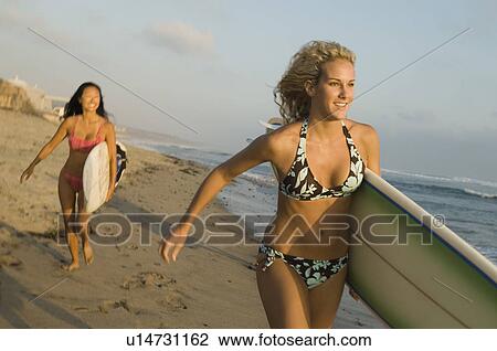 Two female surfers carrying surfboards, running on beach View Large Photo Image Stock Image - Two female surfers carrying surfboards, running on beach. Fotosearch