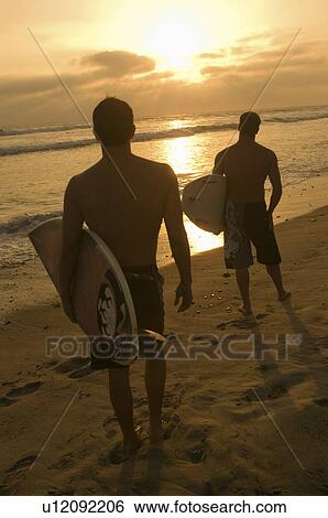 Stock Photograph - Two surfers standing on beach, holding surfboards, watching sunset, back view. Fotosearch