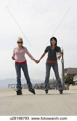 Stock Image - Two women on roller blades holding hands on street. Fotosearch