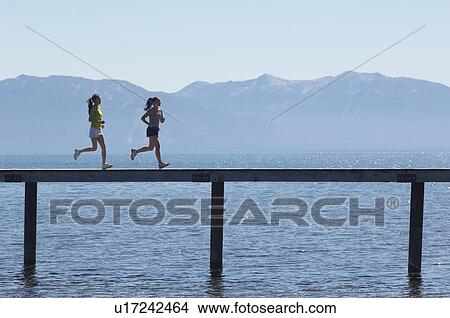 Two women running along a pier with mountains behind, side view View Large Photo Image Picture - Two women running along a pier with mountains behind, side view. Fotosearch