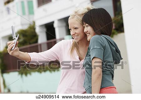 Picture - Two women taking picture with cell phone on city street. Fotosearch