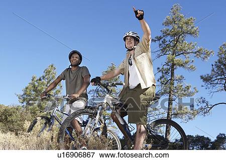 Stock Photo - Two young men on mountain bikes in field, low angle view.. Fotosearch