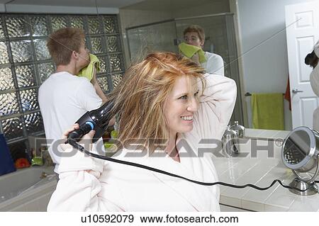 Woman blow-drying hair with boyfriend standing behind in bathroom View Large Photo Image Stock Photo - Woman blow-drying hair with boyfriend standing behind in bathroom. Fotosearch