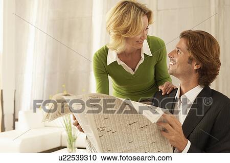 Stock Image - Woman greeting man reading newspaper, indoors. Fotosearch