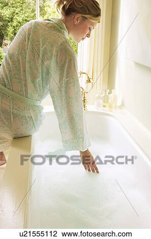 Stock Image - Woman in bathrobe bending down over bathtub filled with bubbles, testing water, back view. Fotosearch