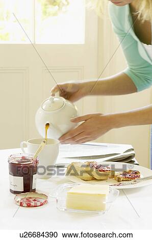 Stock Image - Woman Pouring Tea into cup on dining room table at Breakfast. Fotosearch