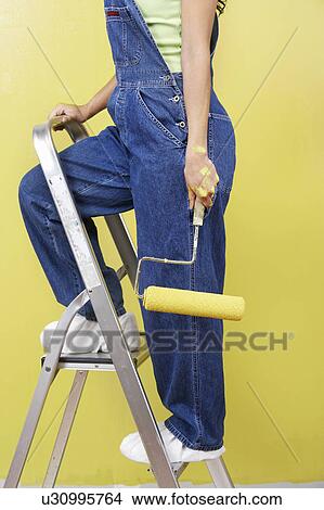 Picture - Woman standing on stepladder holding paint roller, side view, low section. Fotosearch