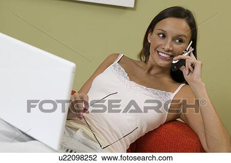 Stock Image - Woman Using Laptop and talking on cell phone on sofa in living room. Fotosearch