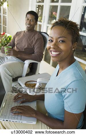 Woman using laptop on porch, husband in background, (portrait) View Large Photo Image Stock Image - Woman using laptop on porch, husband in background, (portrait). Fotosearch