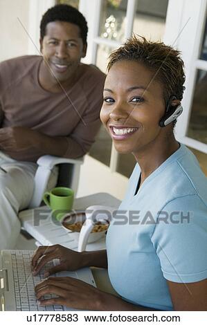 Stock Image - Woman using laptop on porch, husband in background, (portrait). Fotosearch