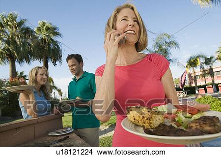Picture - Woman using mobile phone, holding plate of food at family outdoor barbecue.. Fotosearch