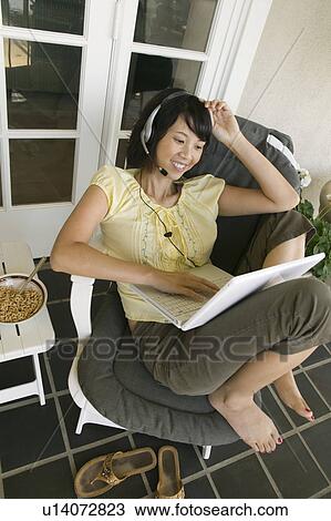 Stock Image - Woman wearing headset using laptop on porch, (elevated view). Fotosearch