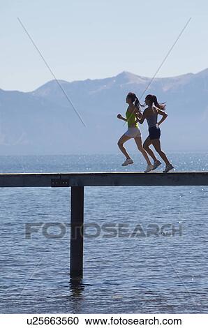 Stock Image - Women running along pier in front of mountains, side view. Fotosearch
