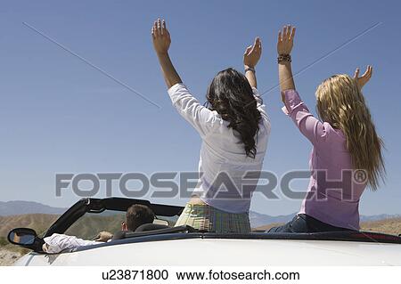 Stock Image - Women with hands raised in the back of a convertible. Fotosearch