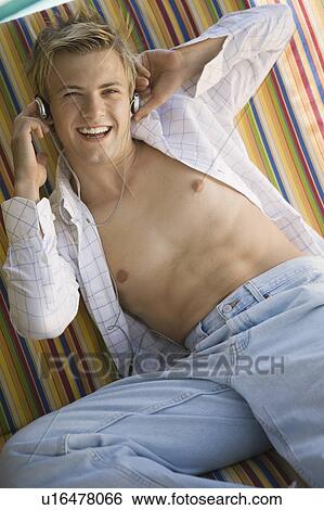 Stock Photograph - Young bare-chested man on deckchair listening to music on headphones, portrait. Fotosearch