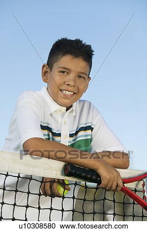 Stock Image - Young boy with tennis racket and ball leaning on tennis net, portrait. Fotosearch