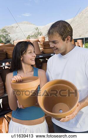 Stock Image - Young couple holding flower pots in outdoors garden centre. Fotosearch