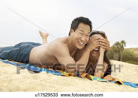 Stock Image - Young couple lying down on beach, laughing, man hiding woman's eyes. Fotosearch
